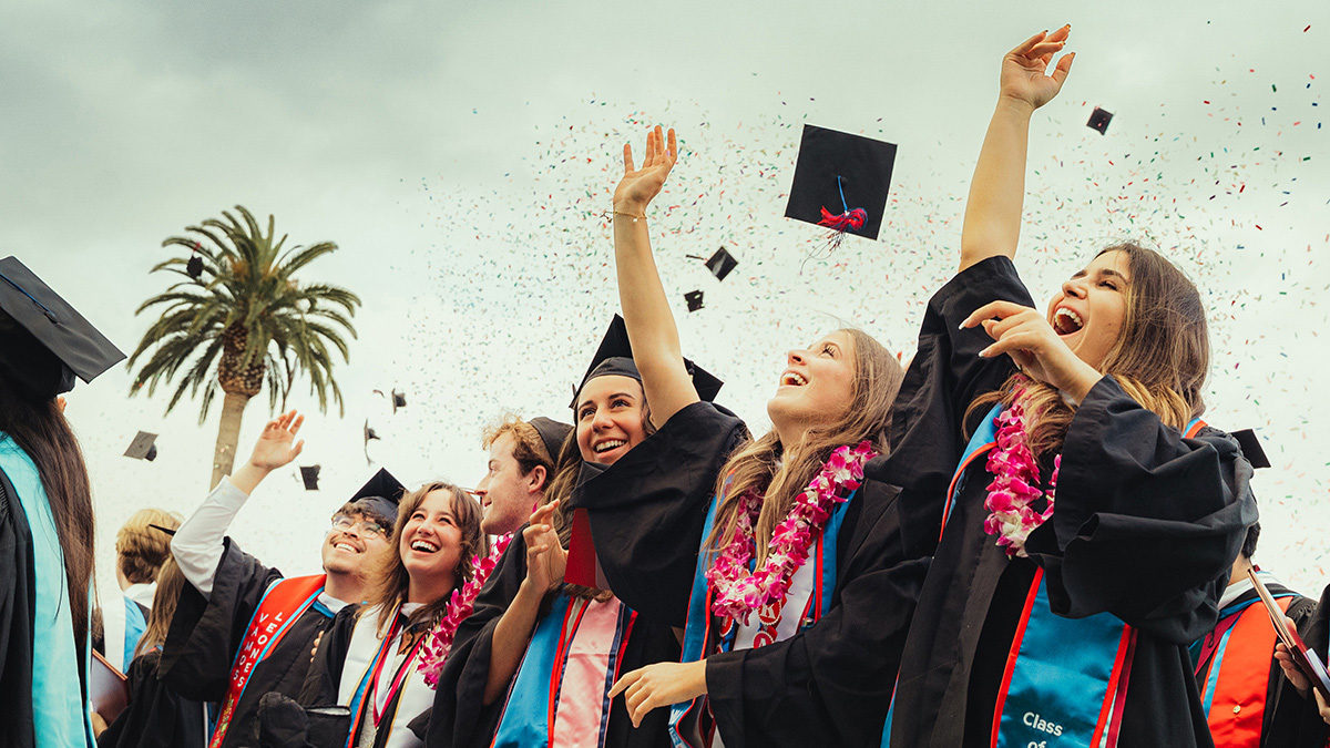 Students celebrating and throwing their graduation caps in the air while confetti floats around them