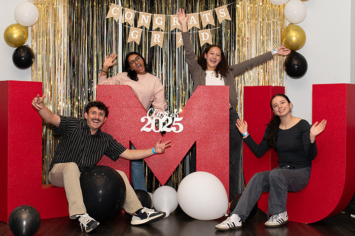 Four students standing and sitting around large L, M, and U letter blocks with balloons and a banner saying CONGRATS GRAD