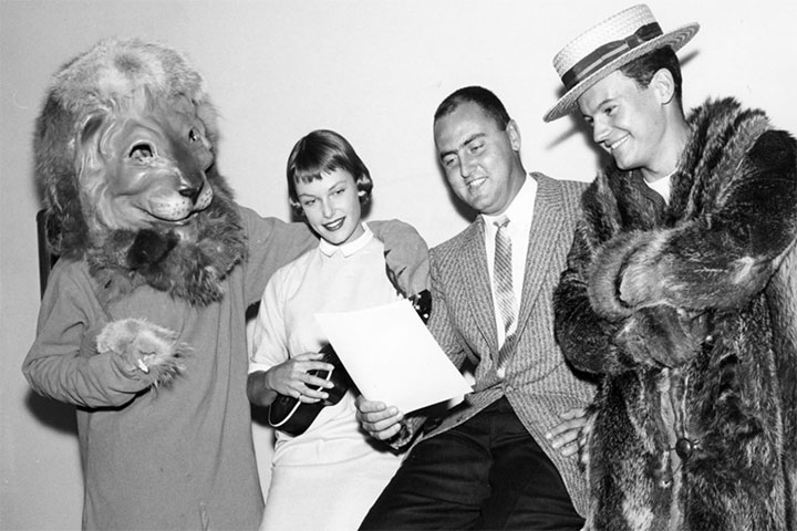 Black and white photo of three students in party attire standing next to a person in an Iggy lion costume